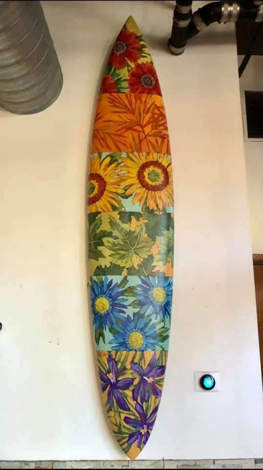 Colorful surfboard with floral designs, laid on a white surface next to a water heater and some pipes. Mark Nilsson ©