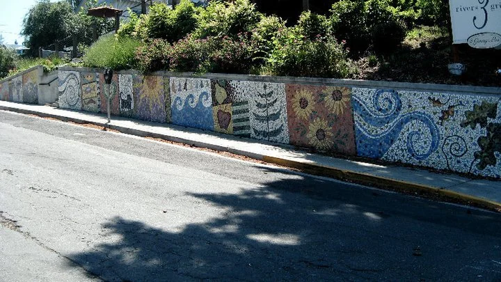 Colorful mosaic mural along a sidewalk featuring various abstract patterns and designs, with a background of greenery and a street.