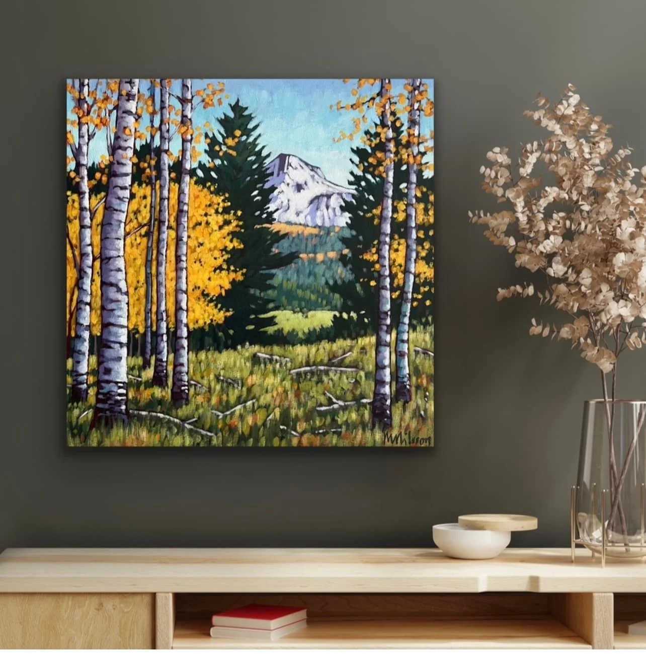 A landscape painting of a forest scene with a mount in the background, framed on a dark wall next to a vase with dried flowers and a small white bowl on a wooden shelf. Mark Nilsson ©