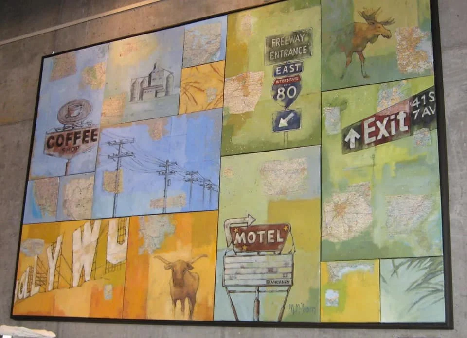 A colorful mural depicting various roadside scenes and signs, including a coffee shop, power lines, a moose, a motel sign, an exit sign, a highway entrance sign, and some trees. Mark Nilsson ©
