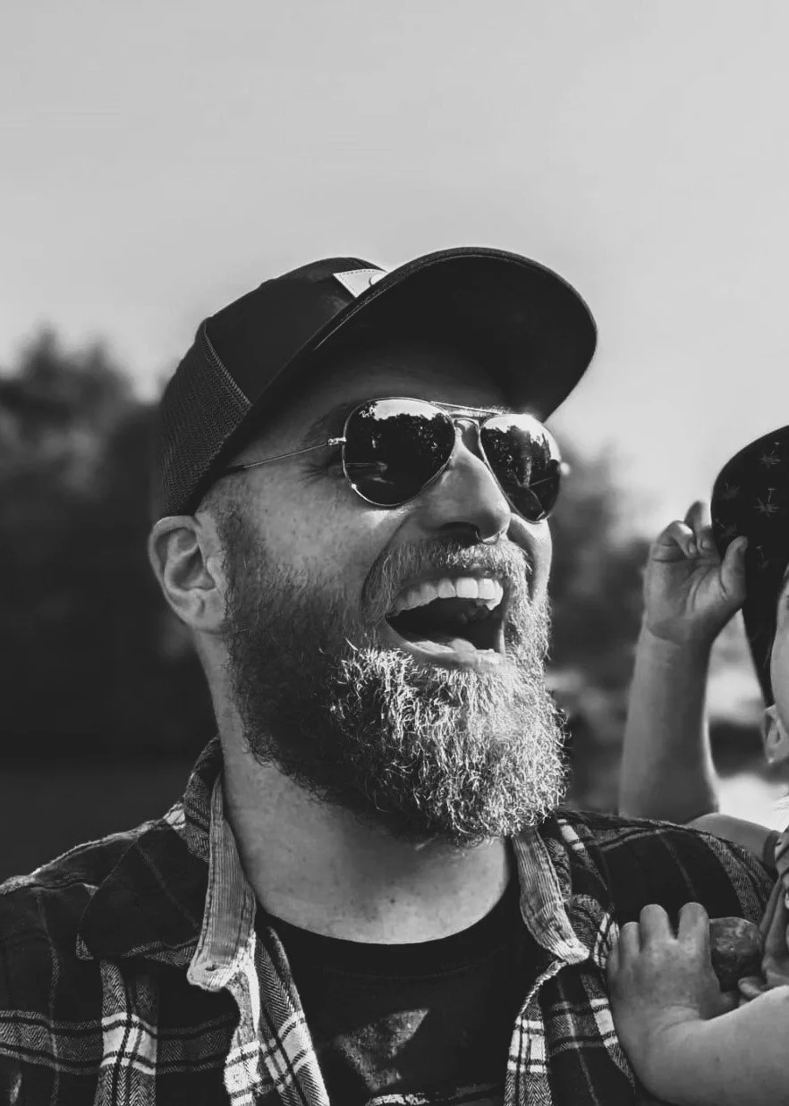Schwarz-weiß-Foto eines lächelnden Mannes mit Bart, Sonnenbrille, Baseballcap und kariertem Hemd, der draußen ist.