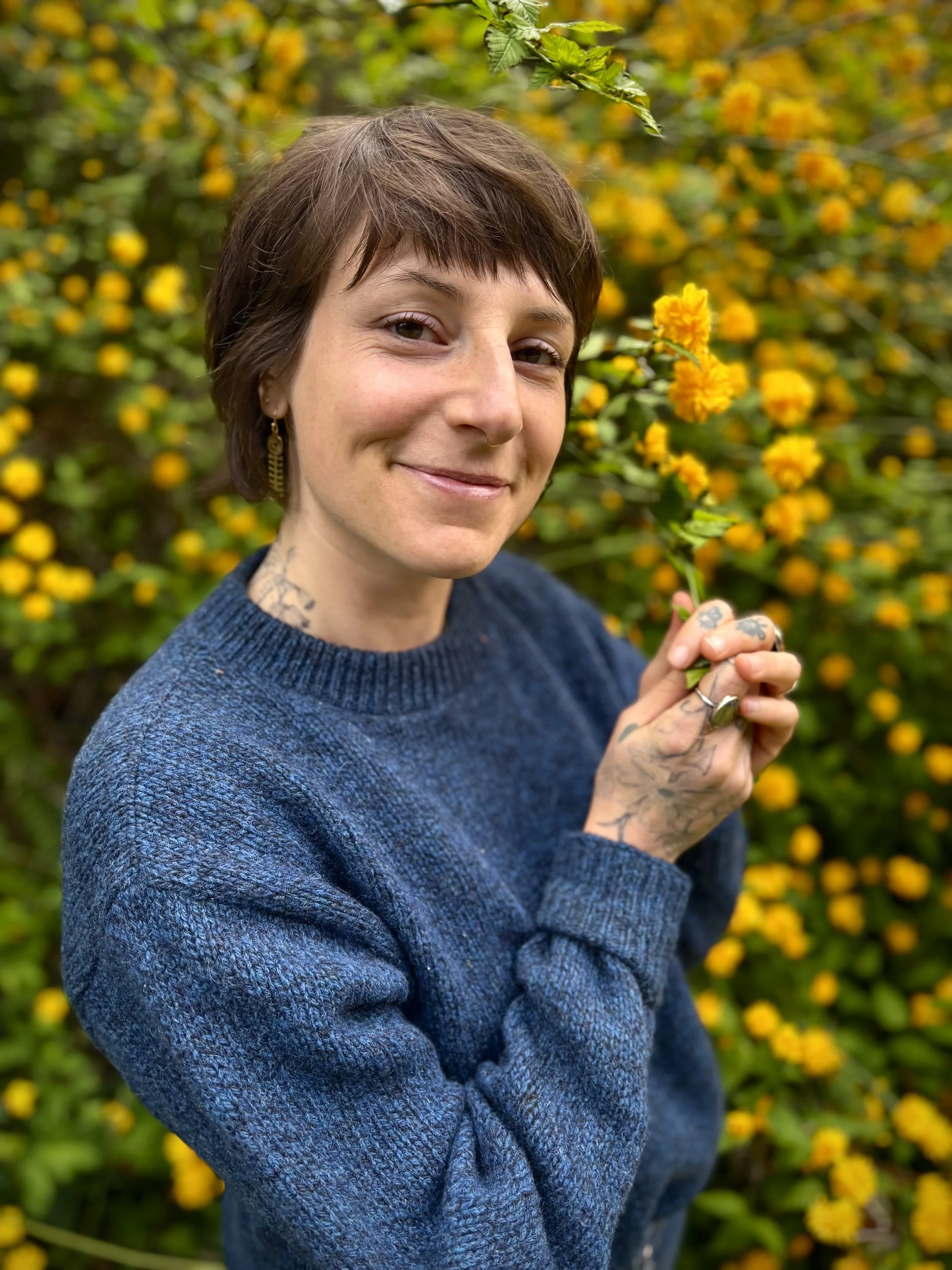 A young woman with short brown hair, tattoos on her hands and neck, and earrings, smiling while holding a yellow flower in front of a bush with yellow and orange flowers.