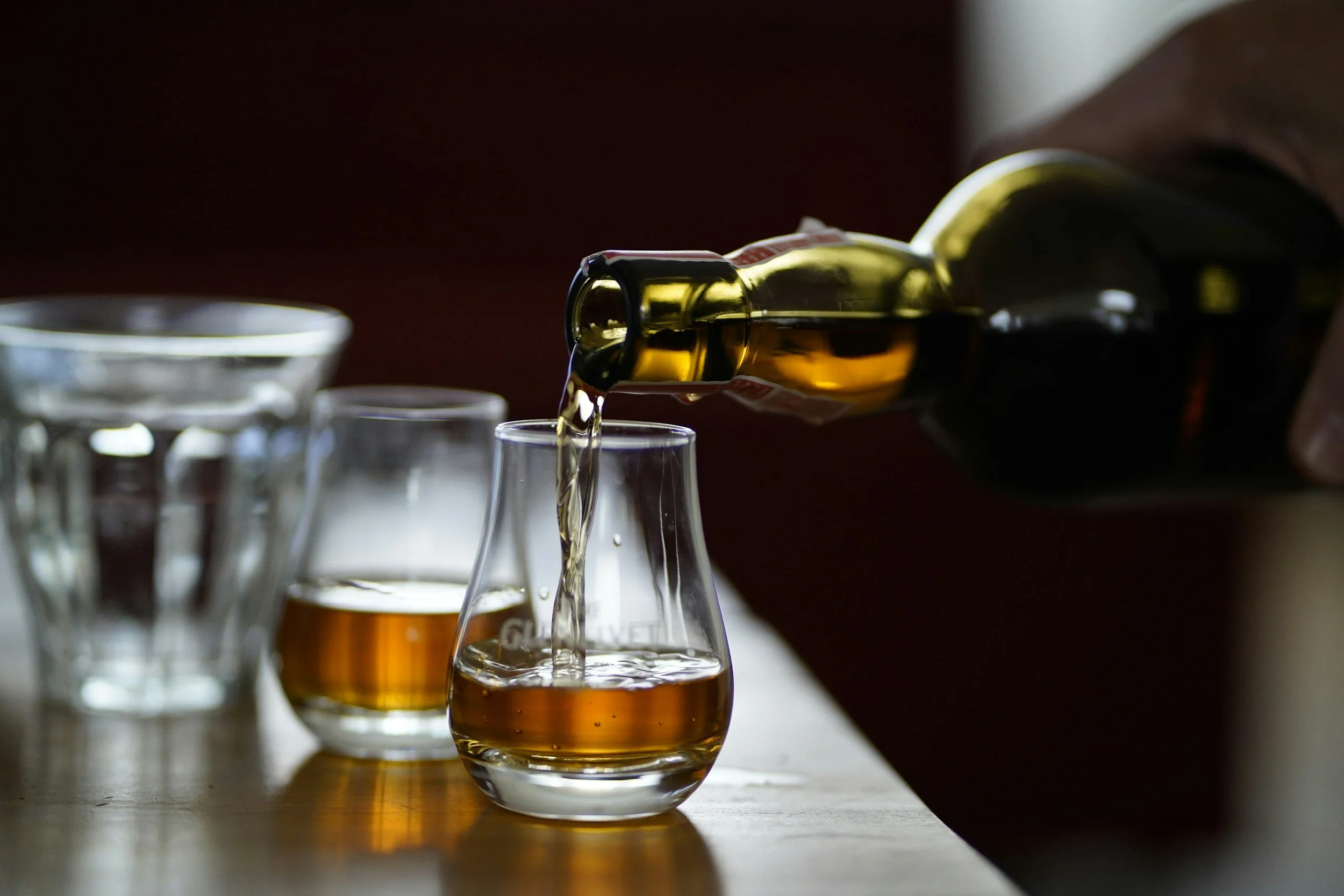A hand pouring whiskey from a bottle into a glass, with additional glasses in the background.
