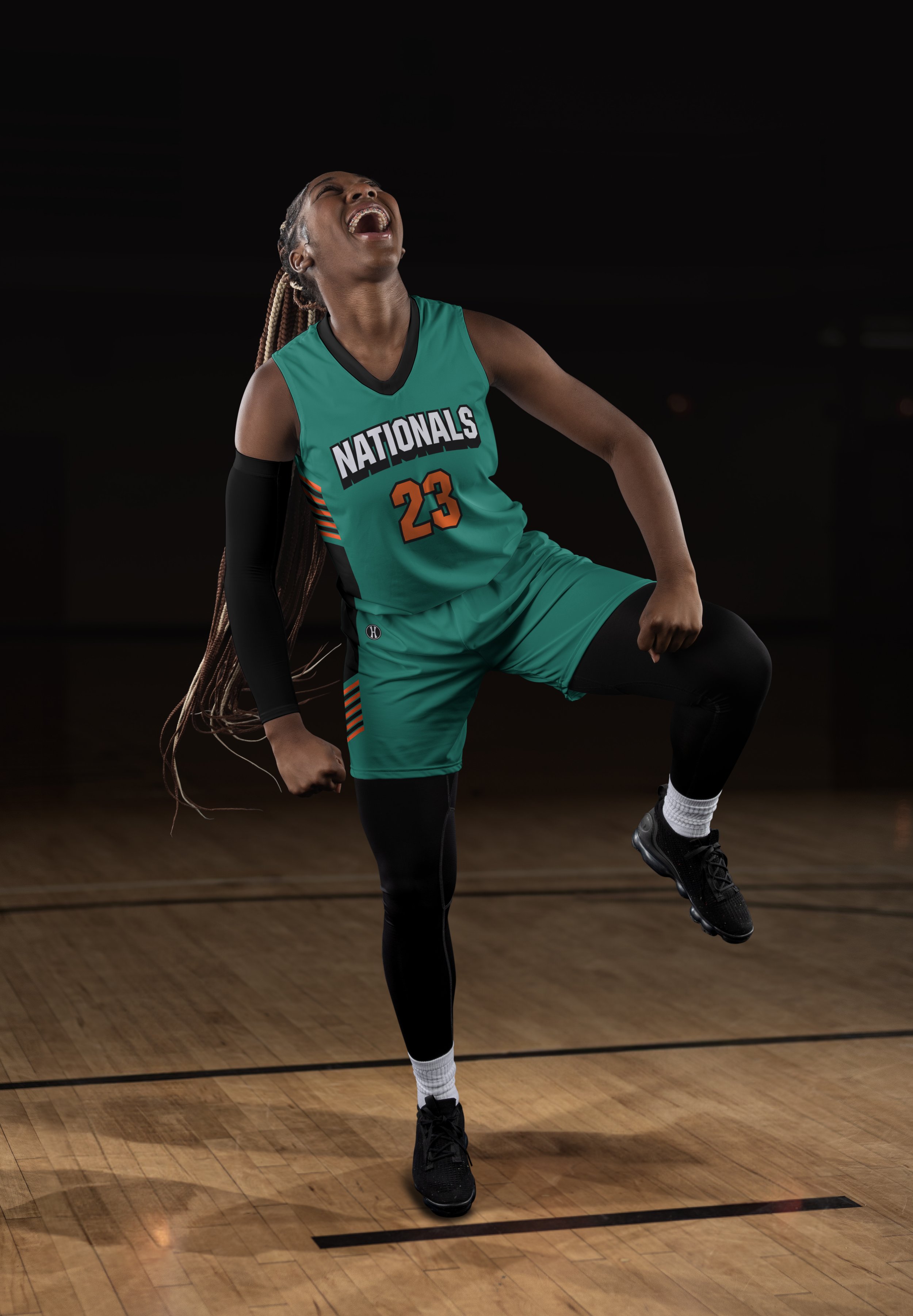 A female basketball player in a teal jersey with the number 23 and the word 'Nationals' is celebrating on a basketball court, with her mouth open in excitement and her fists clenched.