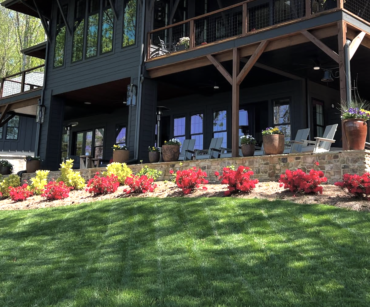A two-story house with a dark exterior, large windows, and wooden balconies. The house has a stone porch with potted flowers and lawn chairs, surrounded by colorful flowers and a well-maintained green lawn.