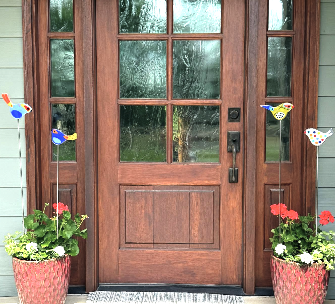 Decorative front door with glass panes, flanked by potted red and white flowers, and colorful bird-shaped garden stakes.