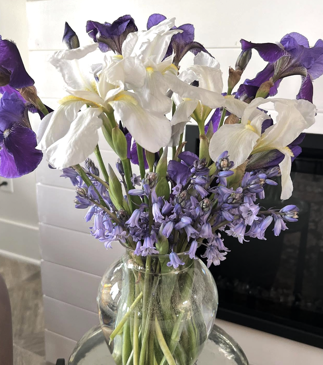 A glass vase filled with white and purple irises and purple hyacinth flowers on a metallic tray.