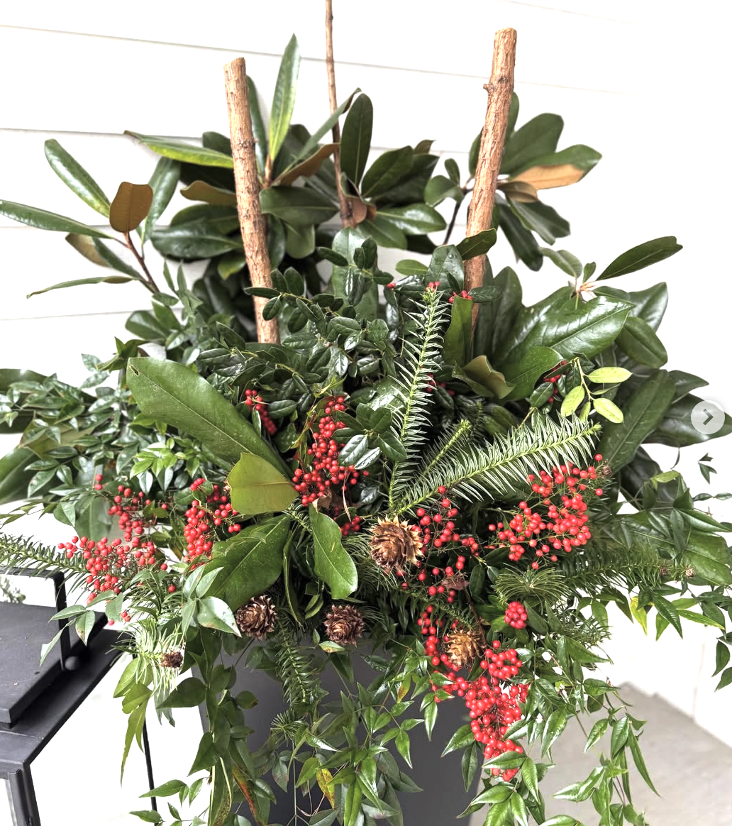 A holiday floral arrangement in a black container with various green leaves, red berries, pinecones, and small decorative sticks, set against a white wall background.