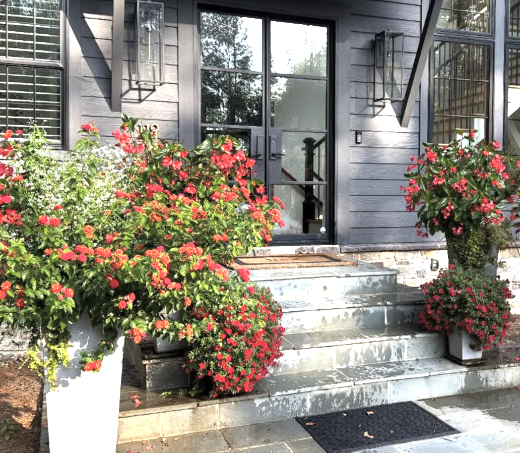 Front porch with stone steps, black door, black lanterns, and potted red and pink flowers.