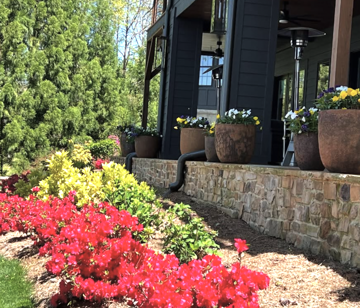 A home with a flower bed filled with pink, yellow, purple, and white flowers in front of a stone foundation. The porch has large flower pots with white and purple flowers, outdoor heaters, and a black exterior wall.