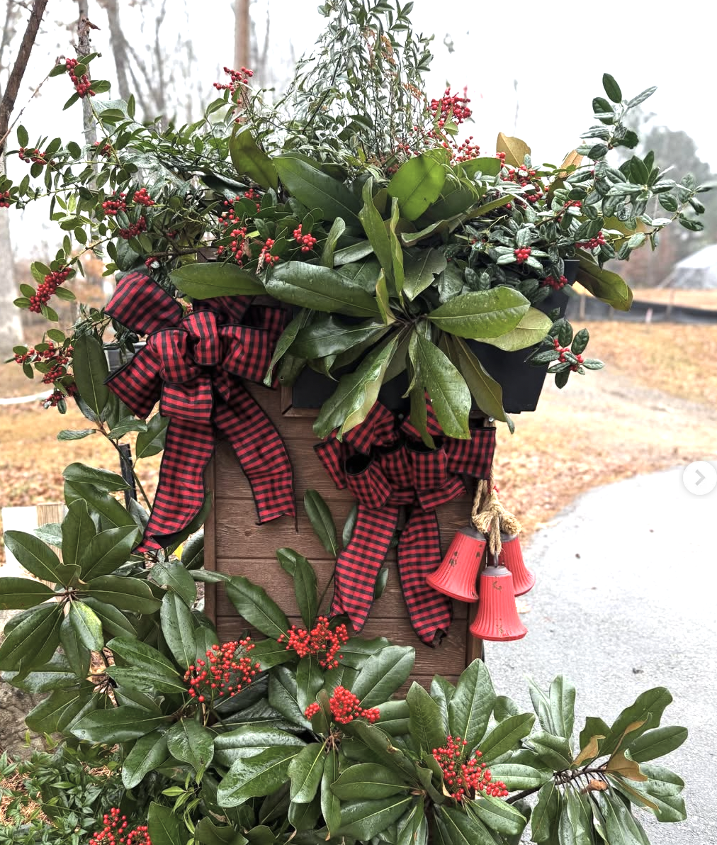 Decorative holiday arrangement with green leaves, red berries, plaid ribbons, and red bell ornaments on a wooden box outside.