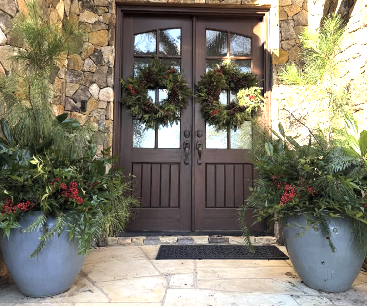 Decorative front door with holiday wreaths, flanked by large potted plants with greenery and red berries.