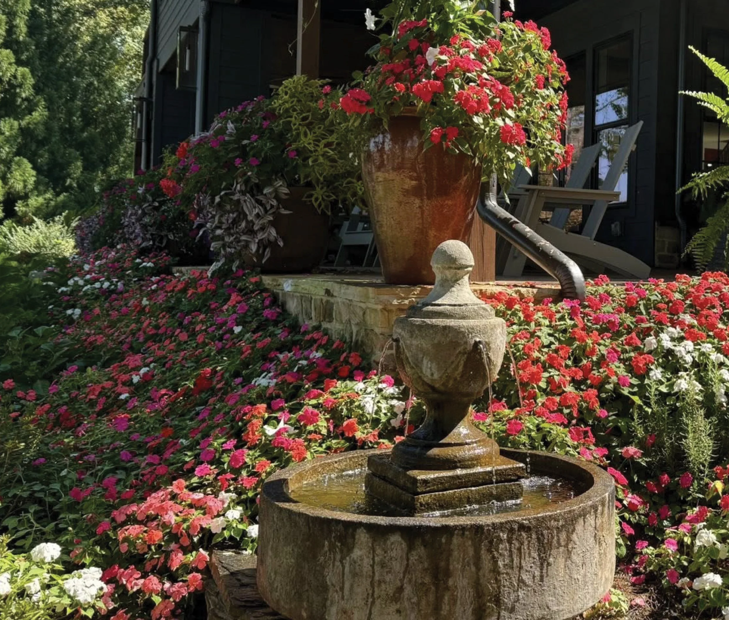 A garden with a stone fountain in the foreground and a porch with potted flowers, including red and pink blooms, in the background.