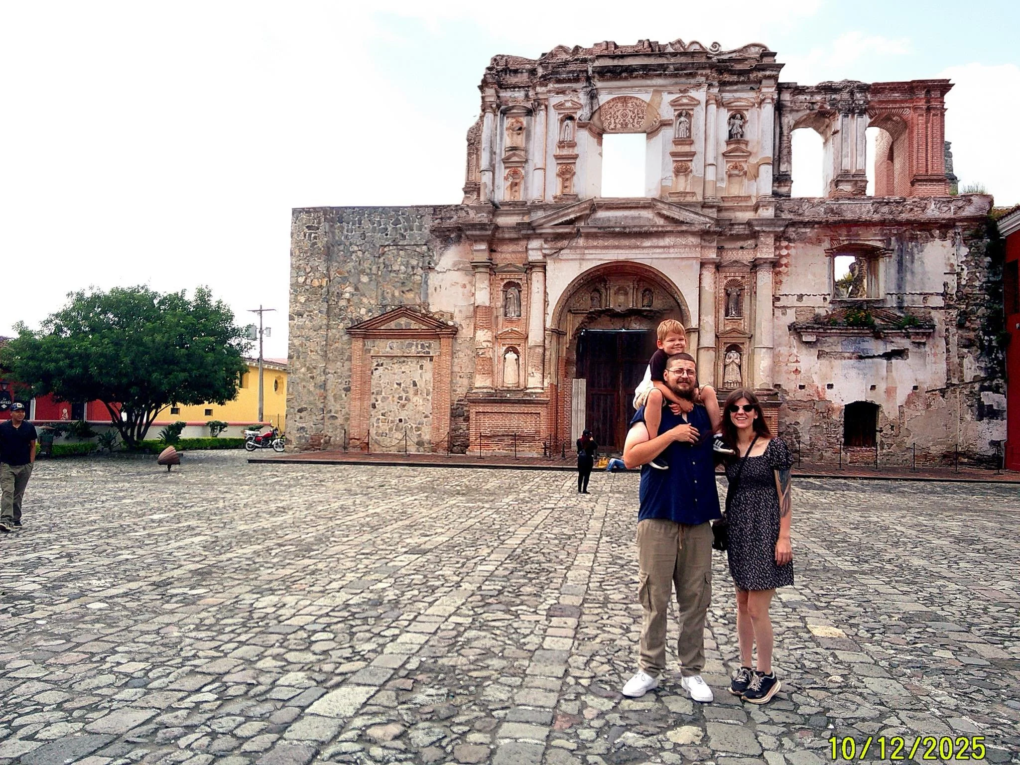 A family of three standing in front of an ancient, weathered stone and brick church or ruins on a cobblestone plaza, with trees and colorful buildings in the background.