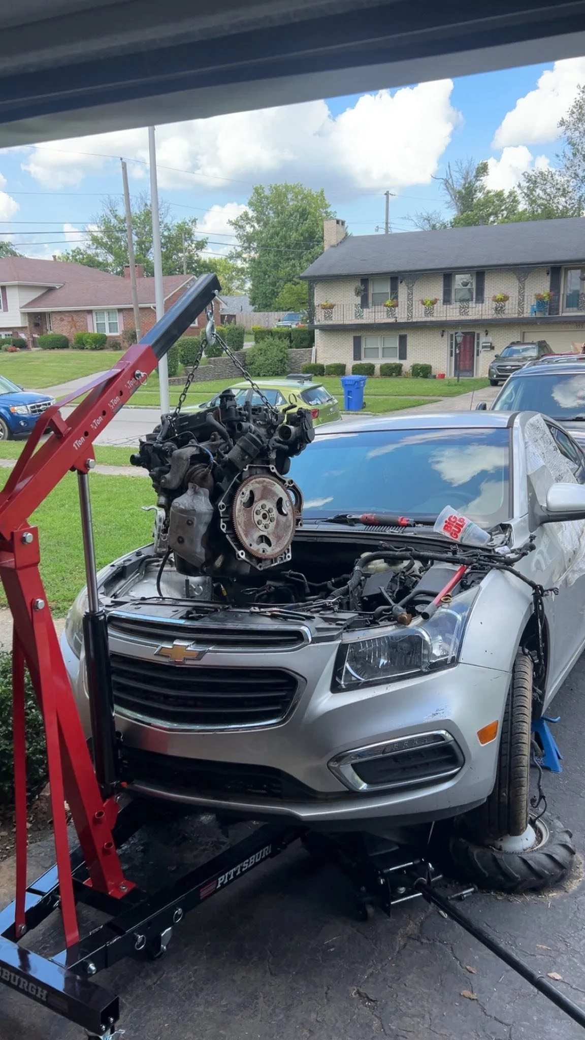 A silver Chevrolet car on a repair lift with an engine hoisted above the open hood, in a residential driveway. The front left tire is removed, and a red engine hoist is holding the engine. The background shows houses, trees, and parked cars on a partly cloudy day.