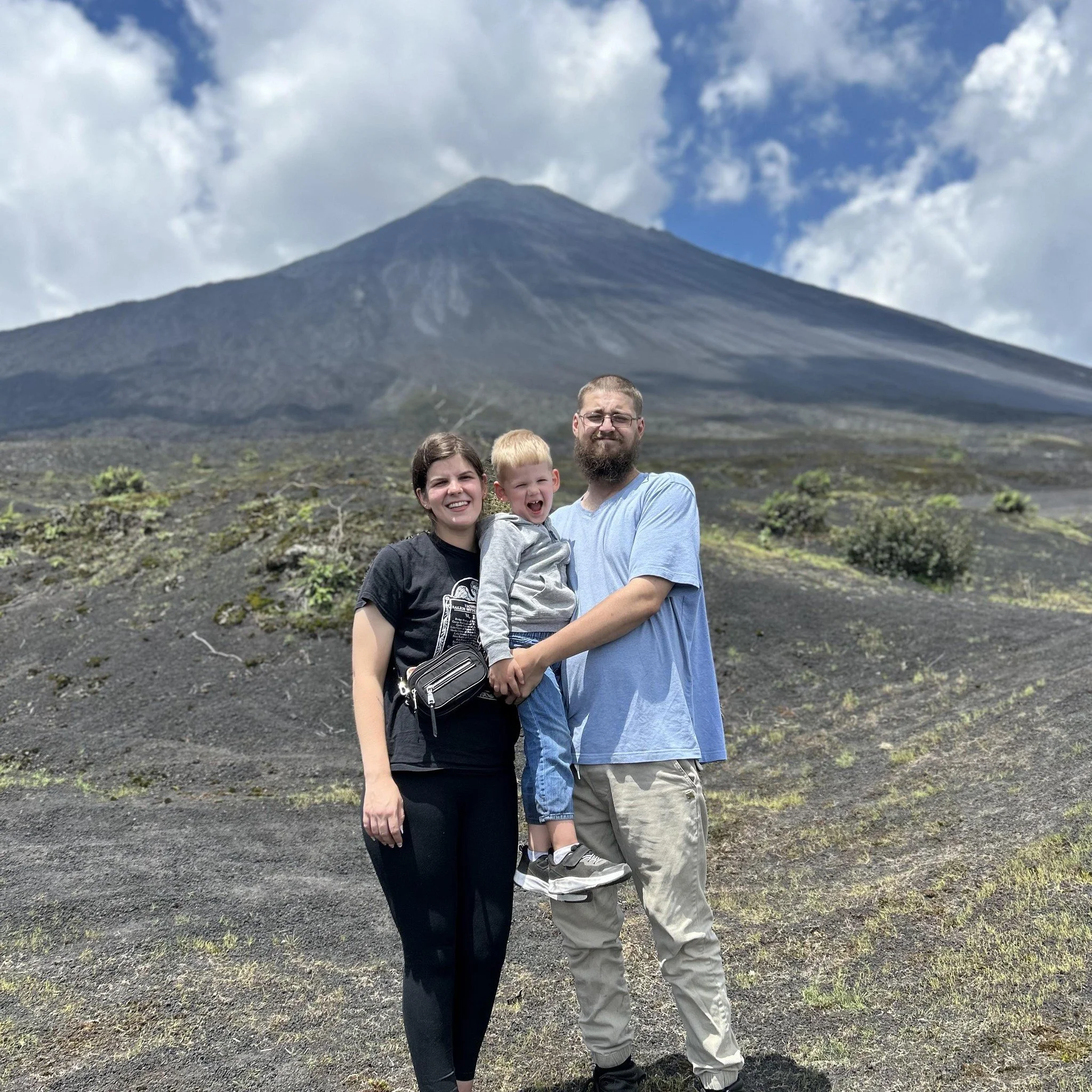 A family of three smiling and standing in front of a large, volcano with a dark slope and a peak, under a partly cloudy sky.