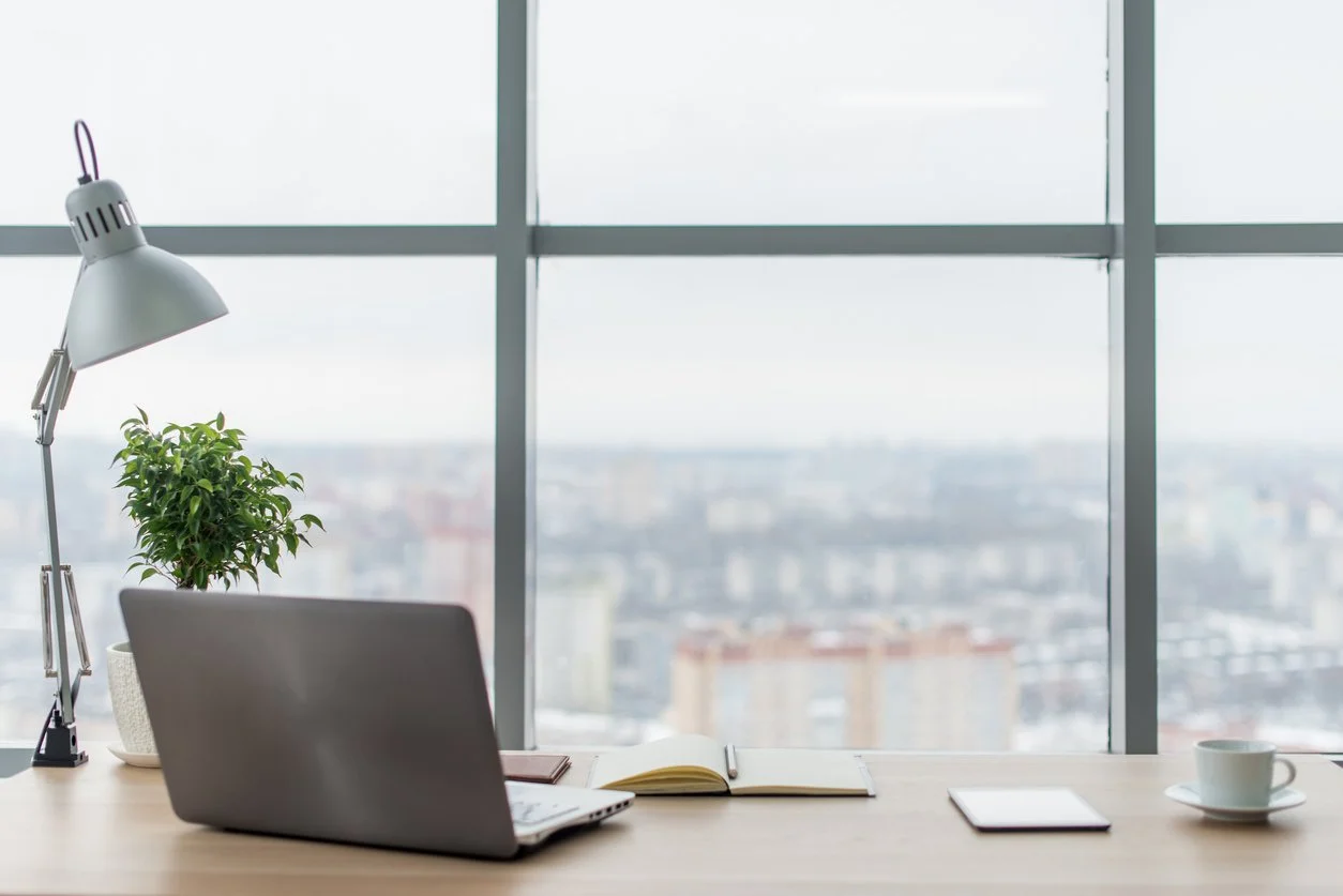 An organized office desk next to a large window overlooking a cityscape, with a laptop, an open notebook, a tablet, a white coffee cup, a potted plant, and a silver adjustable lamp.