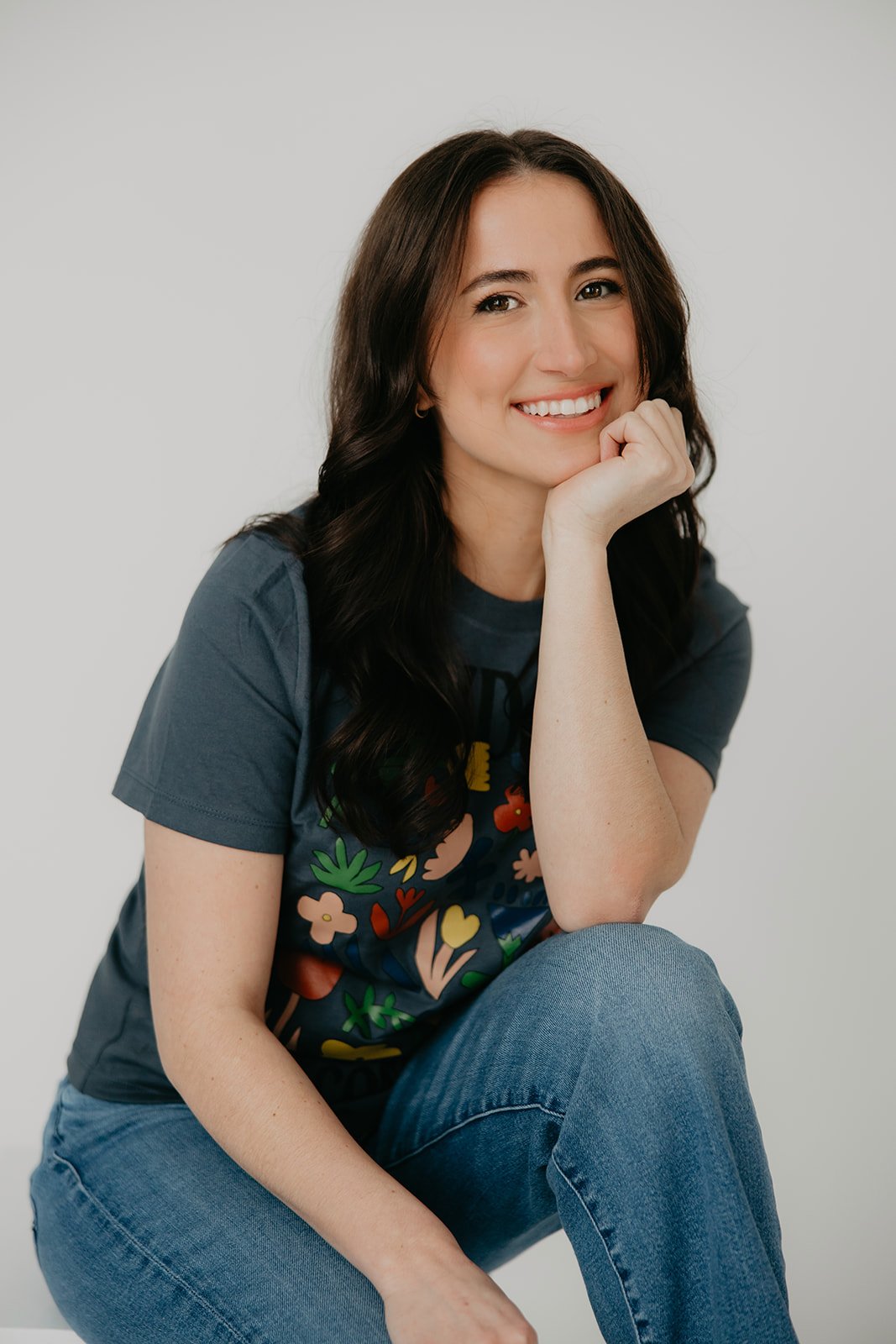 Lauren, a pediatric speech-language pathologist and founder of Shared Spark Pediatric Therapy, smiling while resting her chin on her hand. She is wearing a colorful graphic t-shirt and jeans against a clean white background in Pittsburgh.
