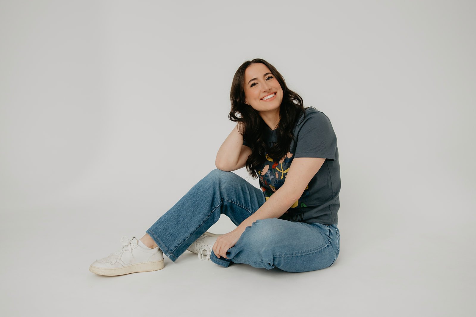 Lauren, a smiling pediatric speech-language pathologist, sits casually on the floor wearing a t-shirt and jeans, representing Shared Spark Pediatric Therapy in Pittsburgh.