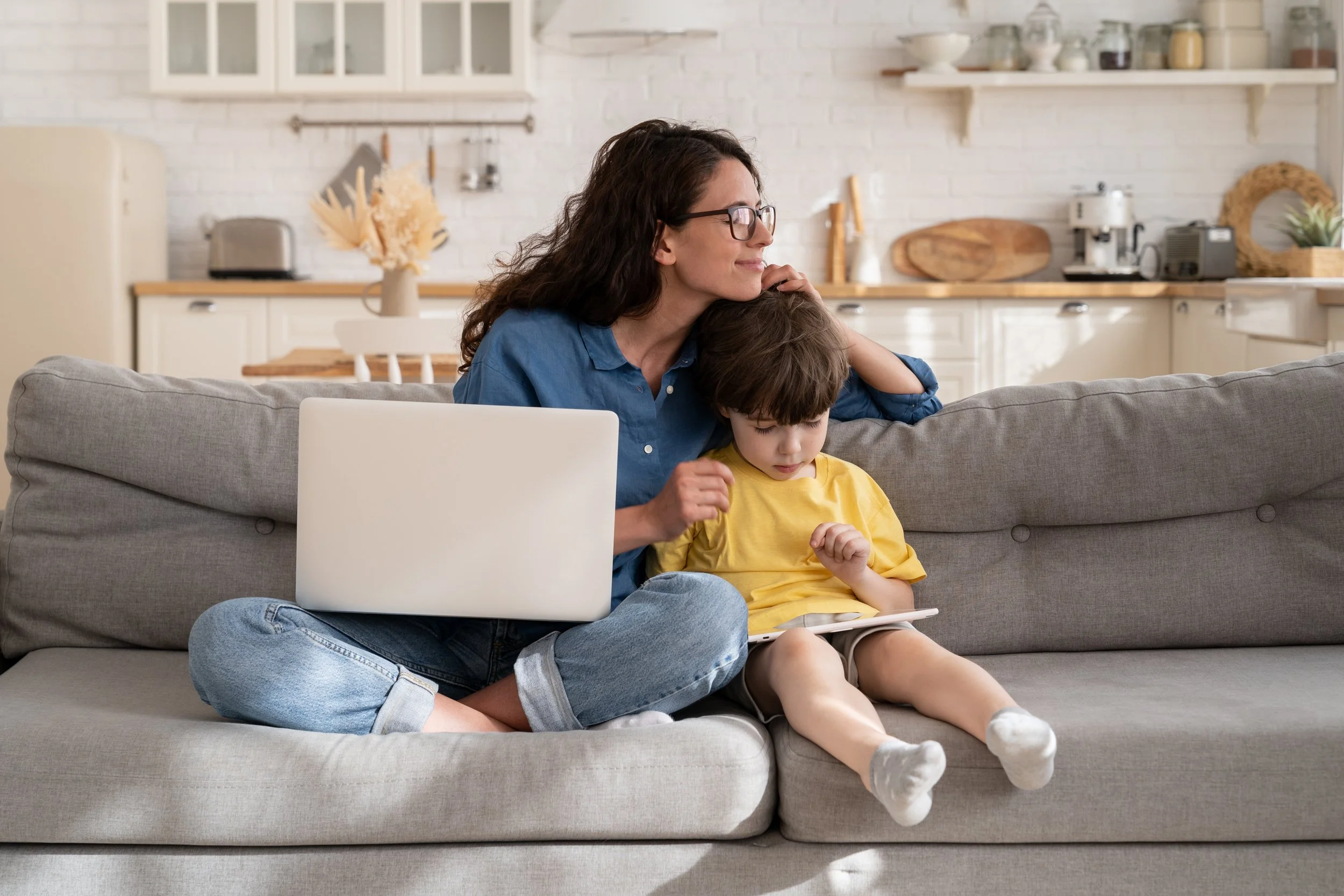 A smiling mother with long curly brown hair and glasses leans in, embracing and looking affectionately at her young son.