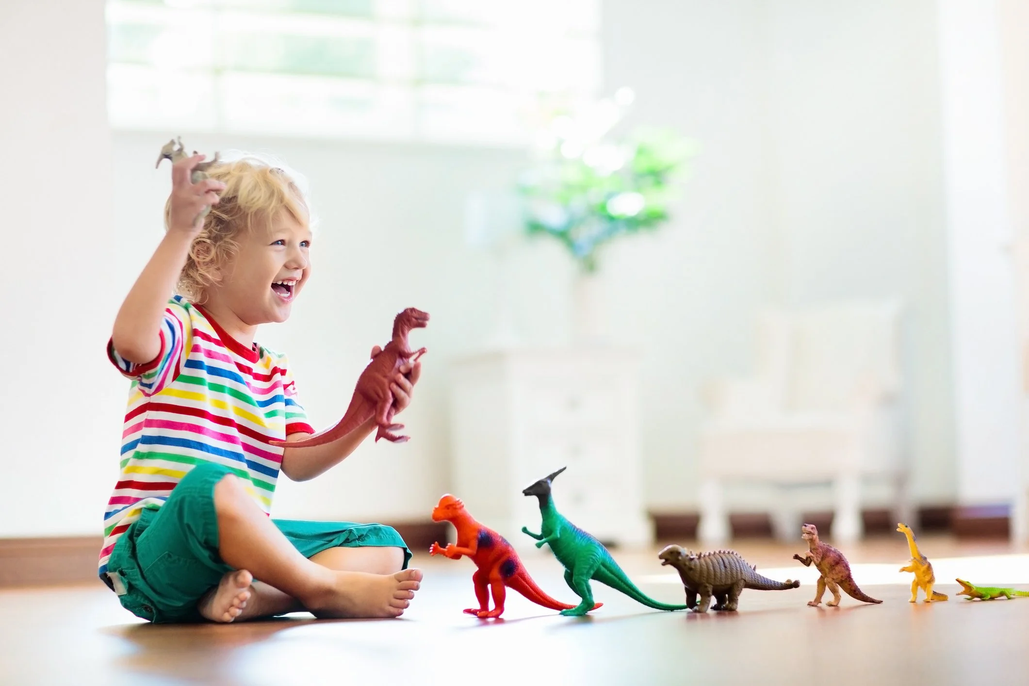 A happy young child with blonde curly hair laughs while playing with toy dinosaurs on the floor during a speech therapy session.