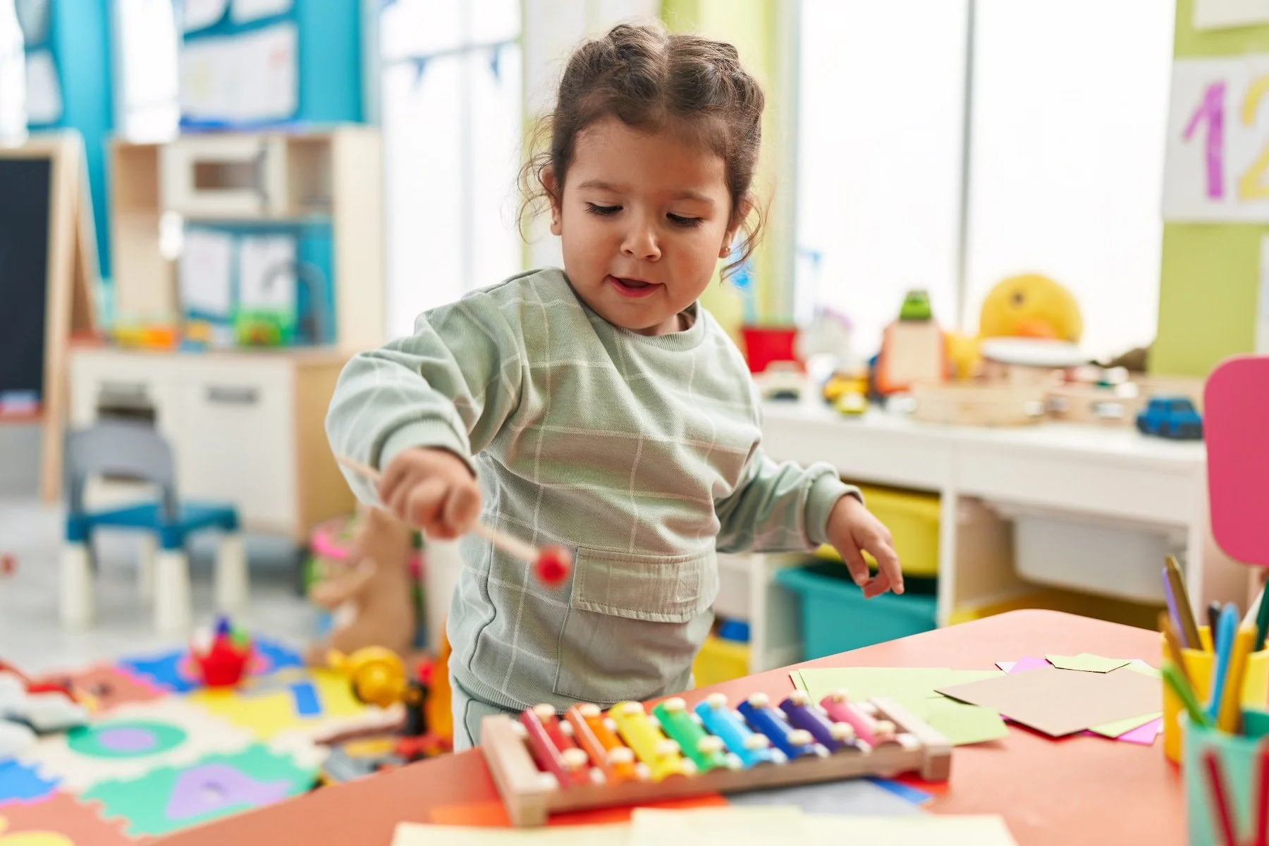 A toddler in a green sweatshirt holds a mallet and plays a rainbow-colored xylophone during speech therapy for late talkers