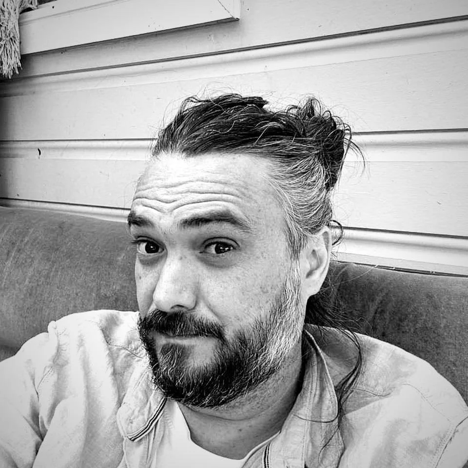 Black and white photo of a man with long hair tied back, a beard, wearing a light-colored shirt, sitting on a bench with a wooden wall background.