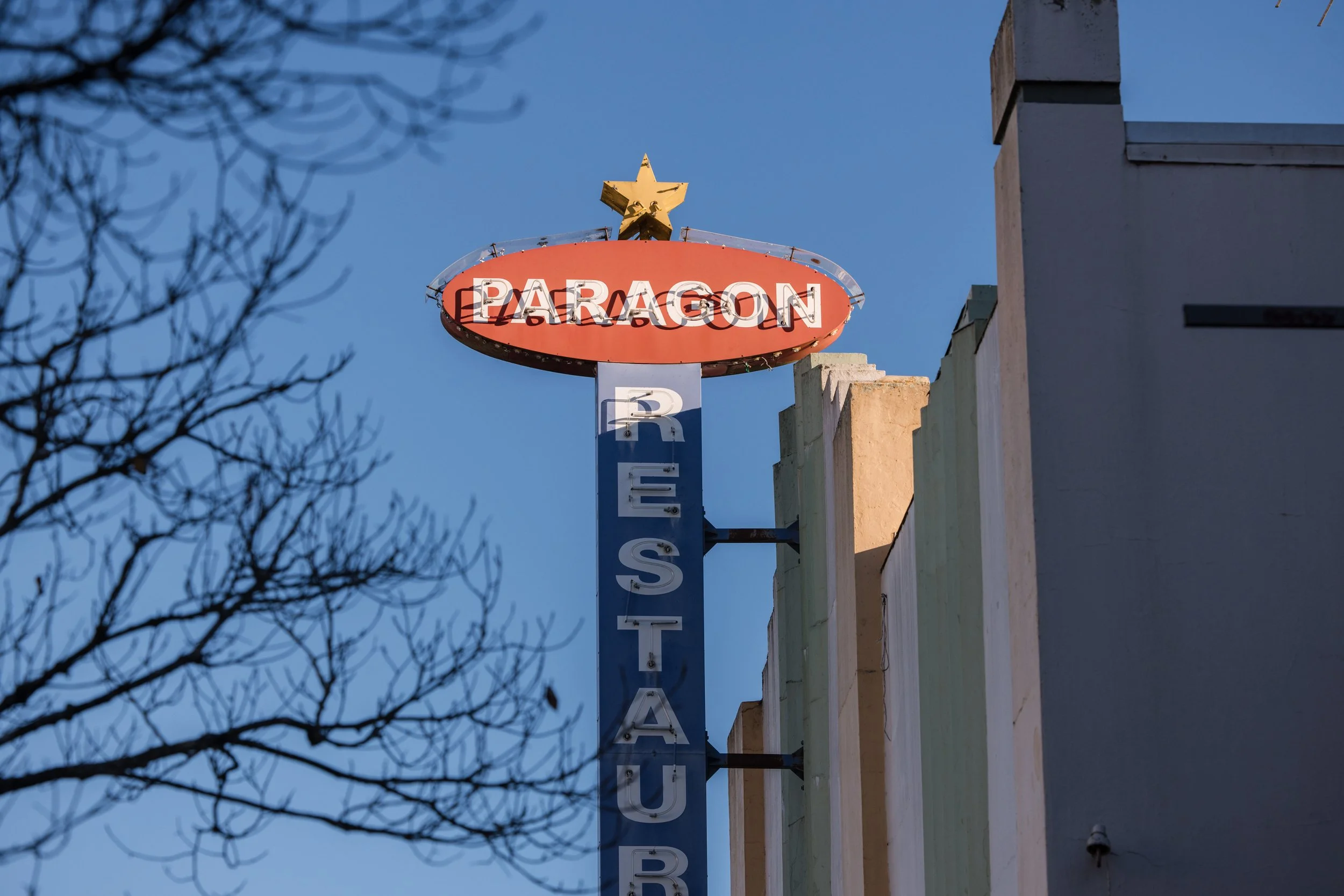 A vintage sign with a gold star on top, displaying the word 'PARAGON' in white letters inside a red oval shape, and partially visible vertical blue and white letters below.