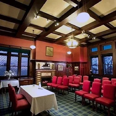 Interior of a room with red chairs arranged in rows, a table with a white tablecloth, wooden ceiling beams, and large windows with diamond-shaped panes.