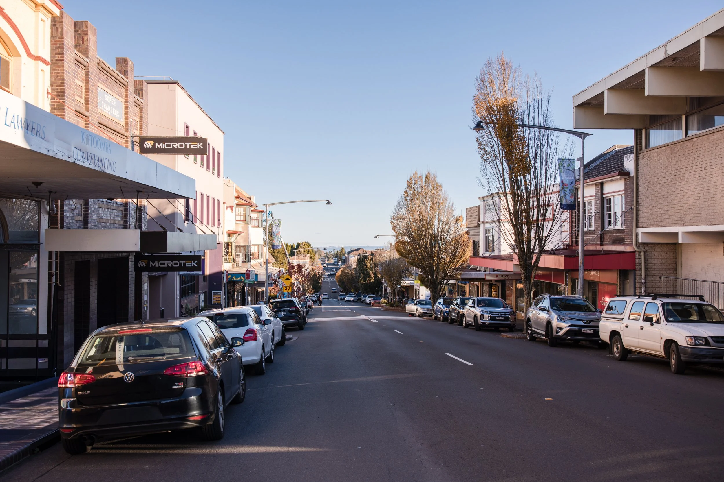 A city street lined with parked cars on both sides, multistory buildings with storefronts, signs, trees with autumn foliage, and a clear blue sky.