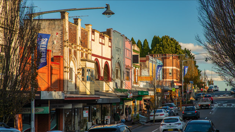 A row of colorful storefronts on a hilly street with parked cars, trees, and pedestrians under a clear blue sky.