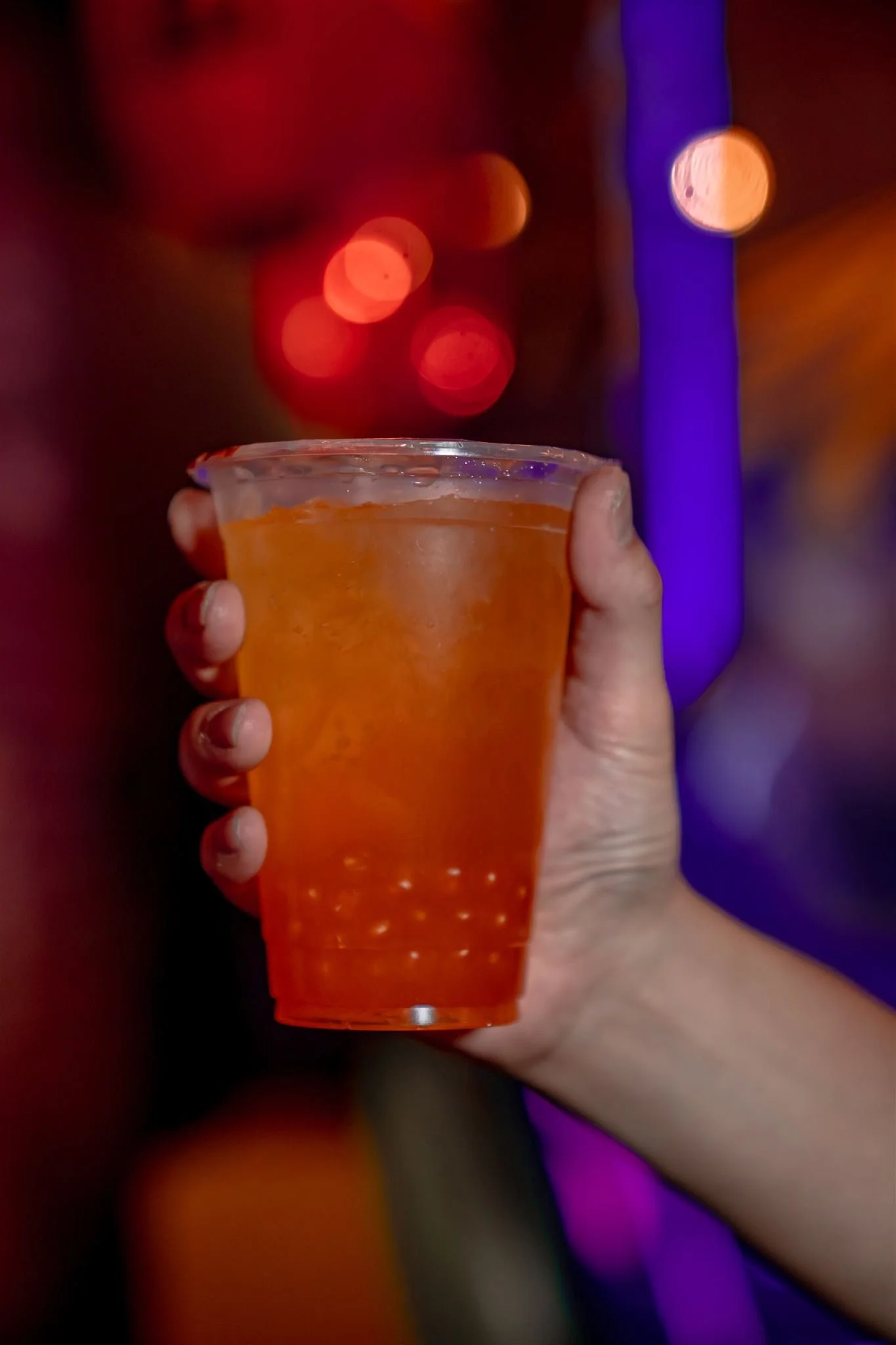 A hand holding a clear plastic cup filled with an orange-colored beverage and ice, against a blurred colorful background with bokeh lights.