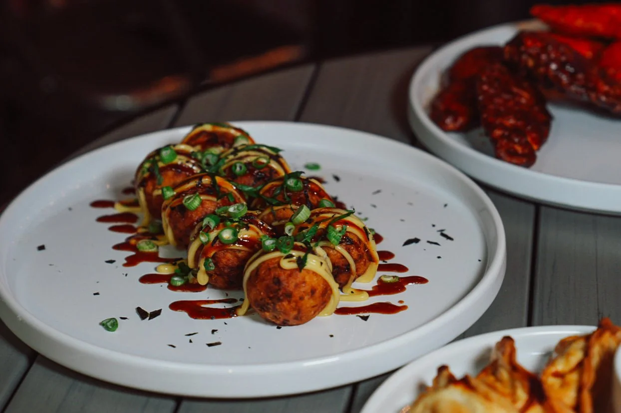 Plate of sushi topped with chopped green onions, drizzled with dark sauce and light mayonnaise, with other Asian dishes in the background.
