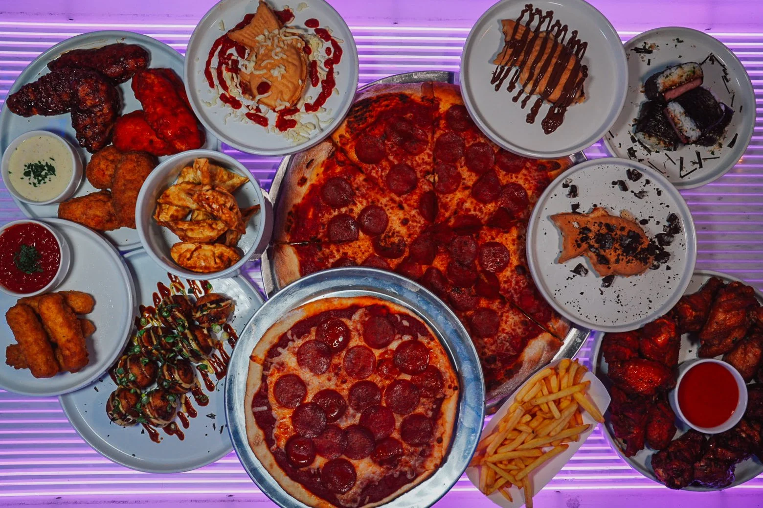 A variety of pizzas, fried chicken wings, sushi, desserts, French fries, and sauces served on multiple plates and bowls on a purple-lit table.