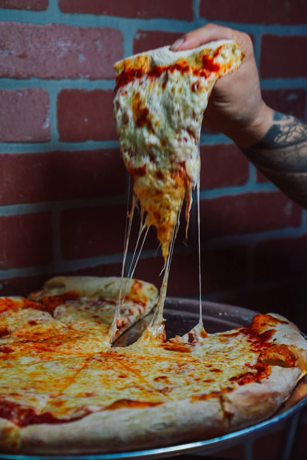 A person lifting a slice of cheese pizza with melted cheese stretching from the slice to the pizza on a tray, against a red brick wall background.