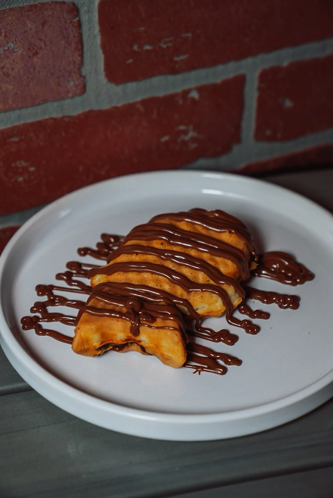 A white plate with three pieces of fried banana drizzled with chocolate sauce, set against a brick background.
