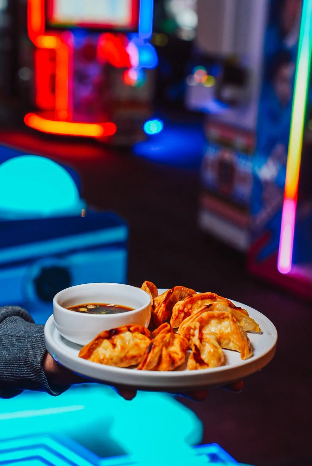 A hand holding a white plate with fried dumplings and a small bowl of dipping sauce in front of a colorful, neon-lit arcade or game room.