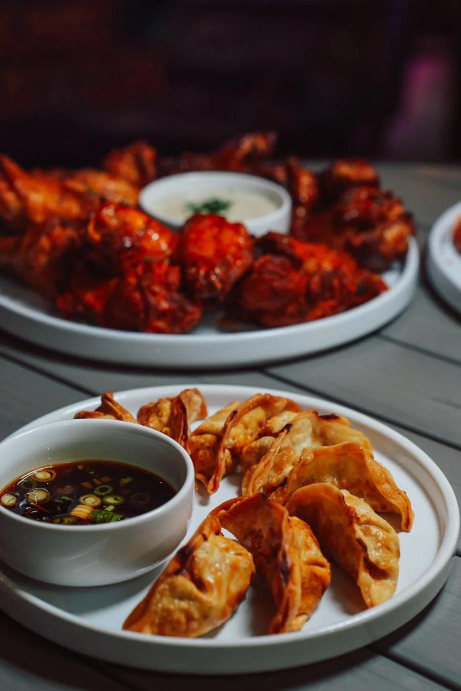 Plate of fried dumplings with soy sauce and green onions, and a plate of spicy chicken wings with dipping sauce, on a wooden table.