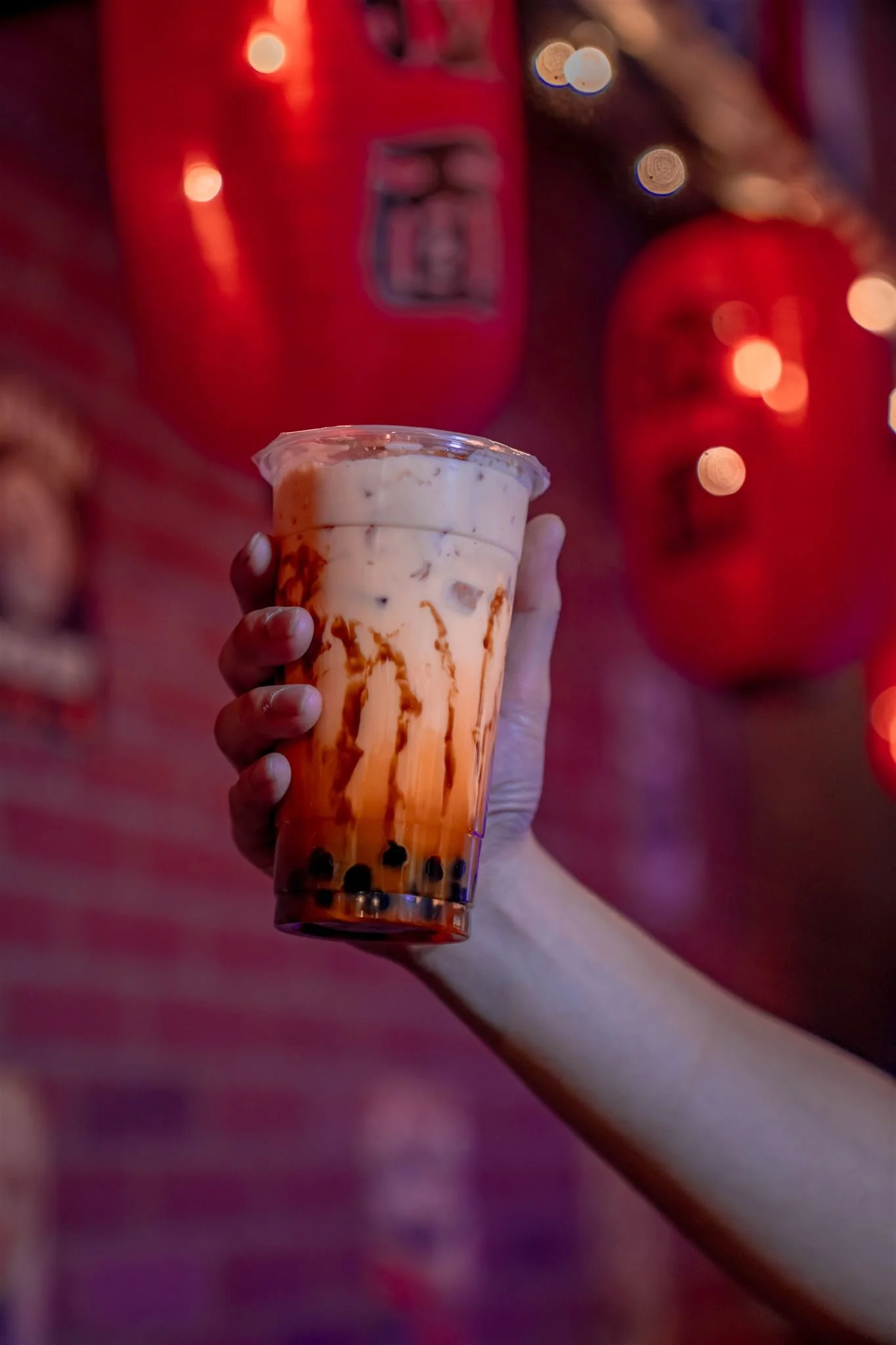 A hand holding a plastic cup of bubble tea with brown sugar syrup, tapioca pearls, and milk. The background includes red lanterns and fairy lights.