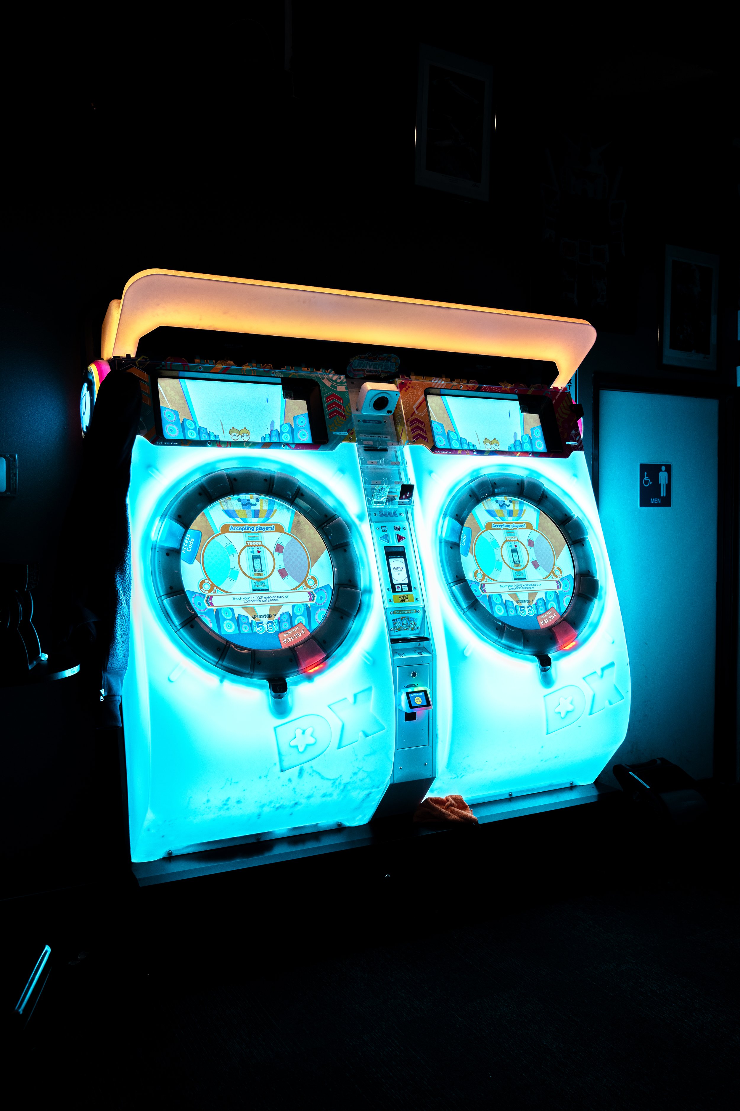 Two illuminated dance arcade machines in a dark room with a bathroom sign in the background.