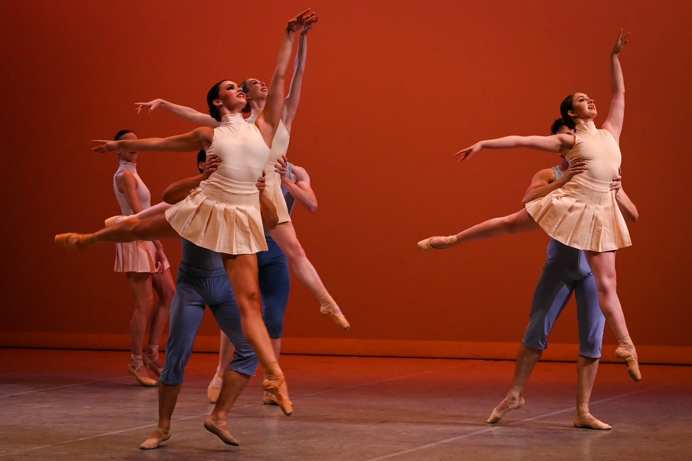 Ballet dancers performing on stage with an orange background, wearing beige and pastel costumes.