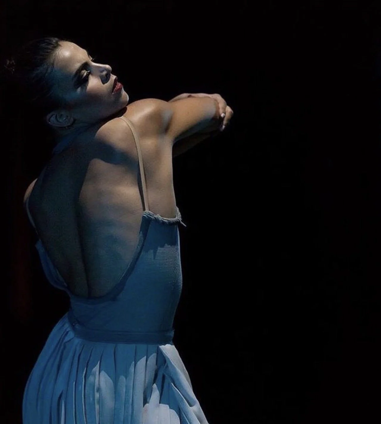 A woman in a dance pose wearing a light-colored dress, with her eyes closed, against a black background.