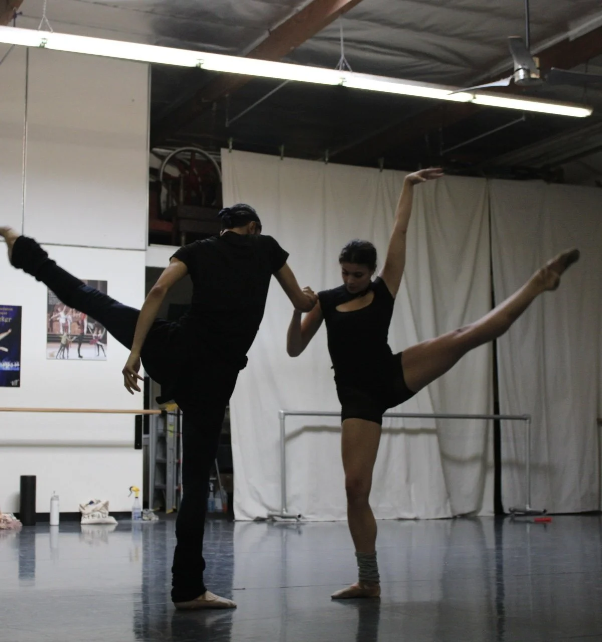 Two ballet dancers practice in a dance studio, one supporting the other, who is extending a leg and arm in a split position.