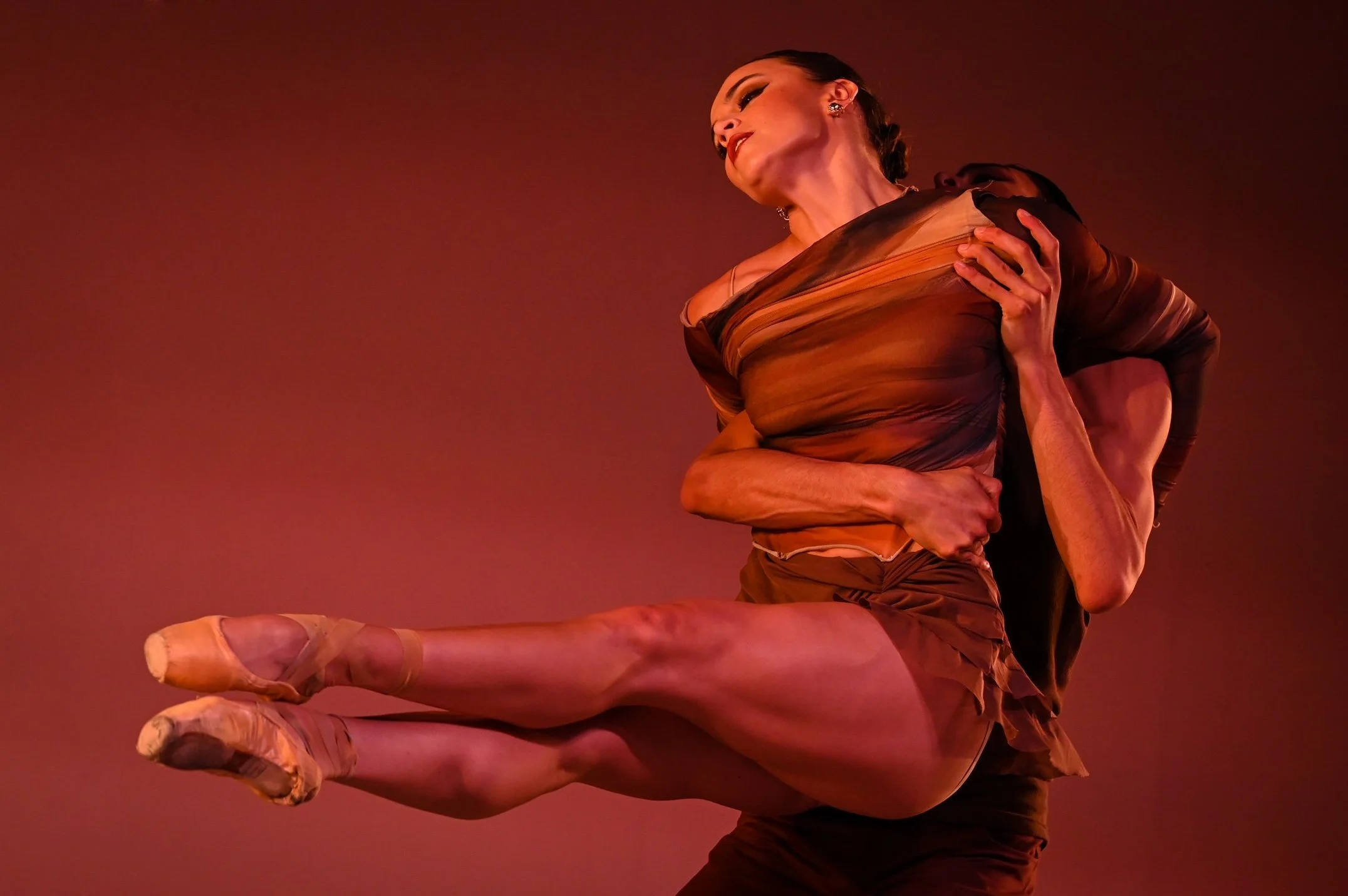 Ballet dancer in mid-air pose, wearing a sheer brown and orange costume, with a plain background.