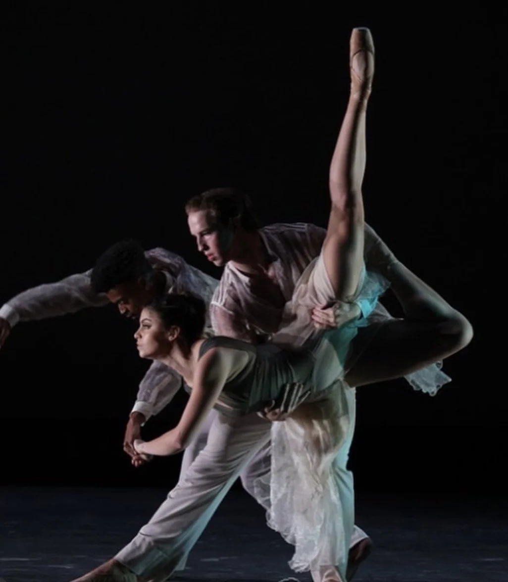 Three ballet dancers performing a pose on stage with a dark background. Two women and one man are engaged in a coordinated dance move.