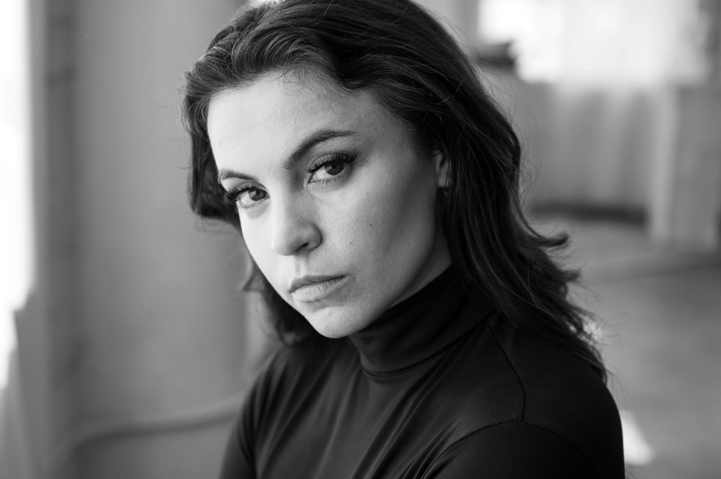 Black and white close-up photo of a woman with dark hair, wearing a high-neck top, looking at the camera with a serious expression.