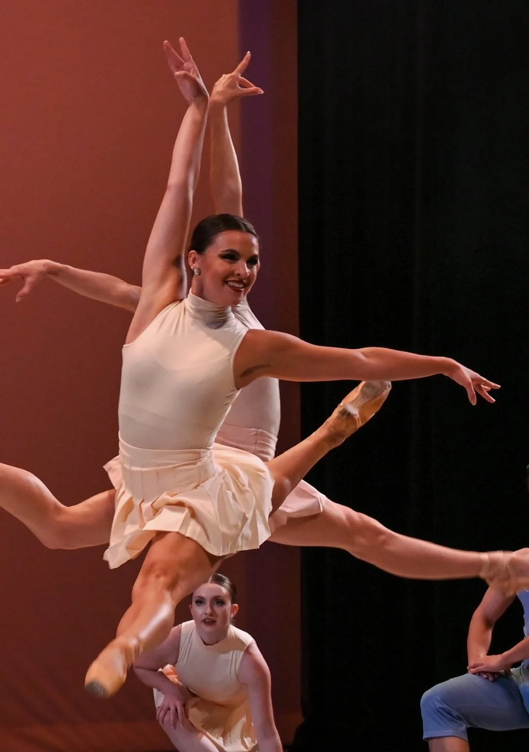 Three female dancers in cream-colored costumes performing an acrobatic routine on stage with a reddish background.