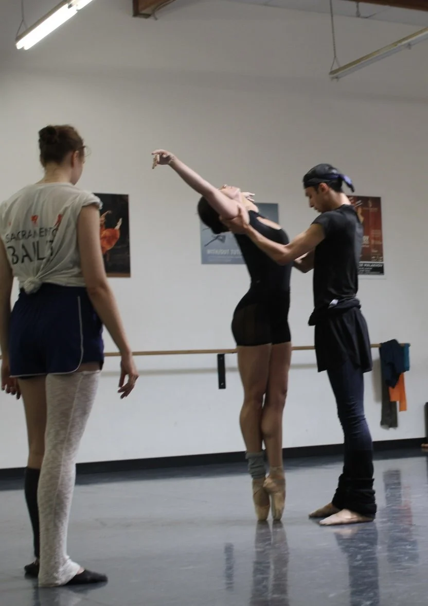 Three ballet dancers in a dance studio practicing. One dancer is en pointe, another is being assisted by a trainer, and the third stands nearby observing.