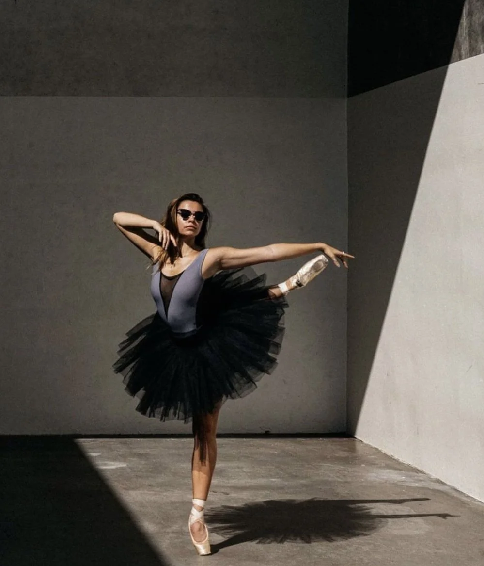 A ballet dancer wearing sunglasses, a black tutu, and pointe shoes performs a pose in a minimalist space with shadowed walls.