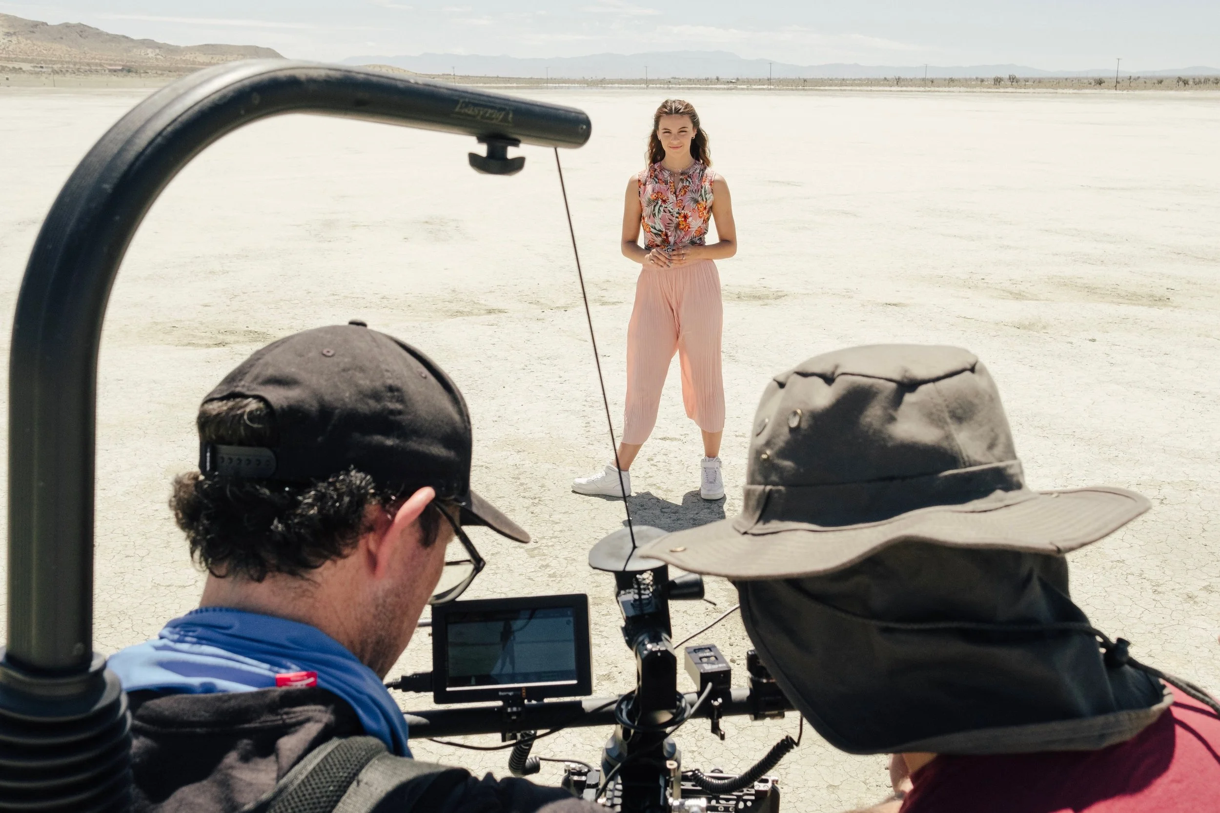 A woman standing on a dry, cracked desert landscape during a photoshoot, with two people filming her, one with a camera and the other with a hat and equipment.
